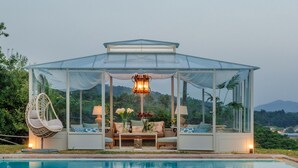 Un pabellón de cristal con una piscina al frente y montañas al fondo.