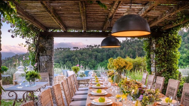 Un espacio de comedor al aire libre con sillas y mesas de mimbre, dispuesto bajo una pérgola de madera con luces colgantes.