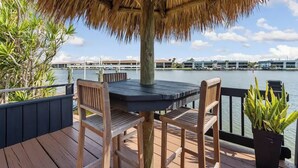 Un pontile in legno con tetto di paglia, tavolo e sedie, affaccia su un corso d'acqua con edifici sullo sfondo.
