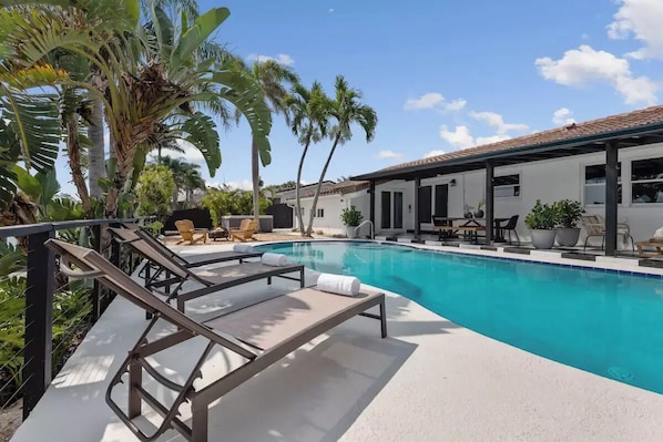 A swimming pool with lounge chairs, palm trees, and a covered patio area.