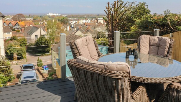 A balcony with wicker furniture and a glass table, overlooking a residential area with houses and a white building in the distance. - The Outlook (Minehead)