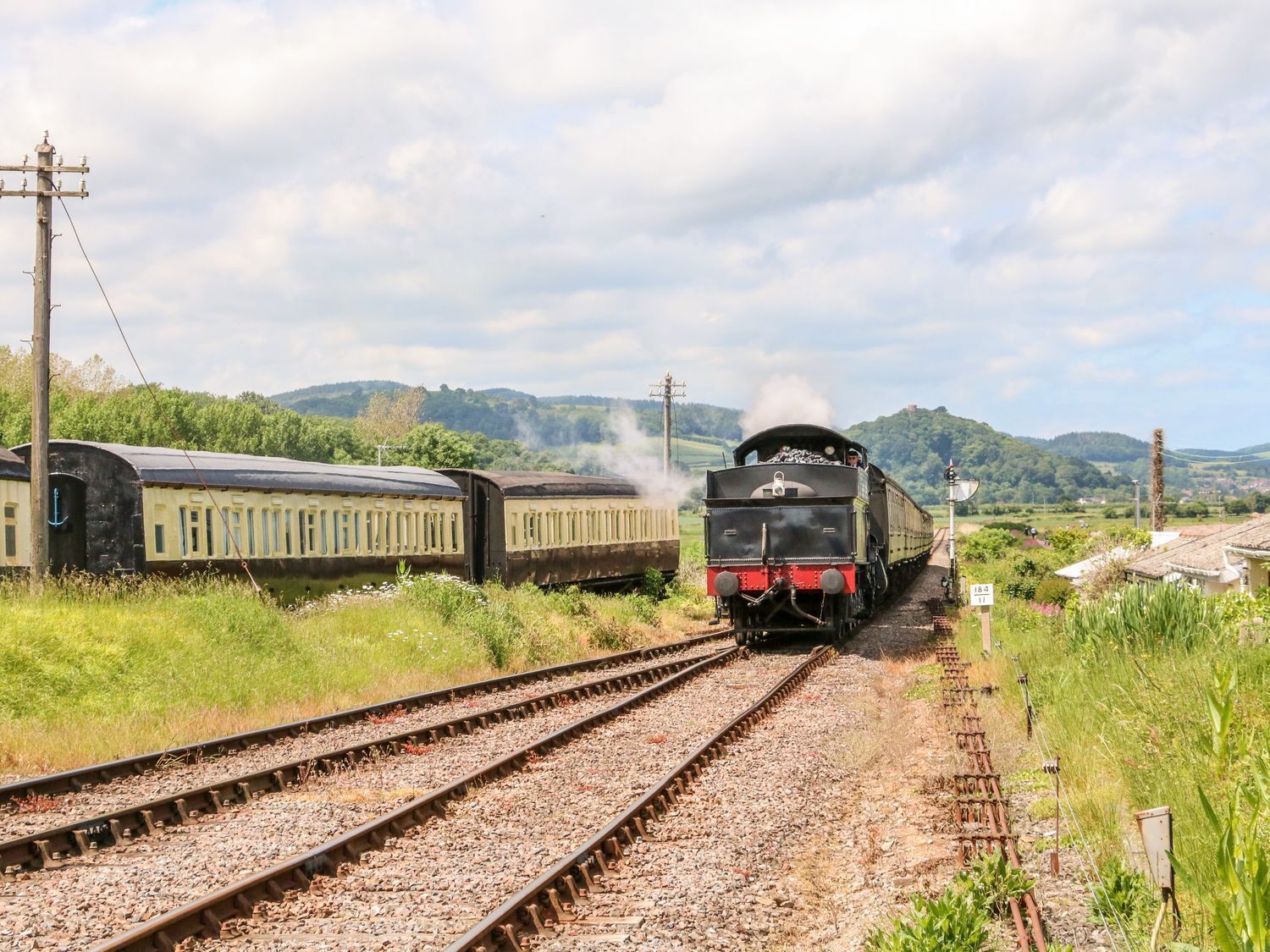 A steam locomotive pulling passenger cars on a railway track with a rural landscape in the background.