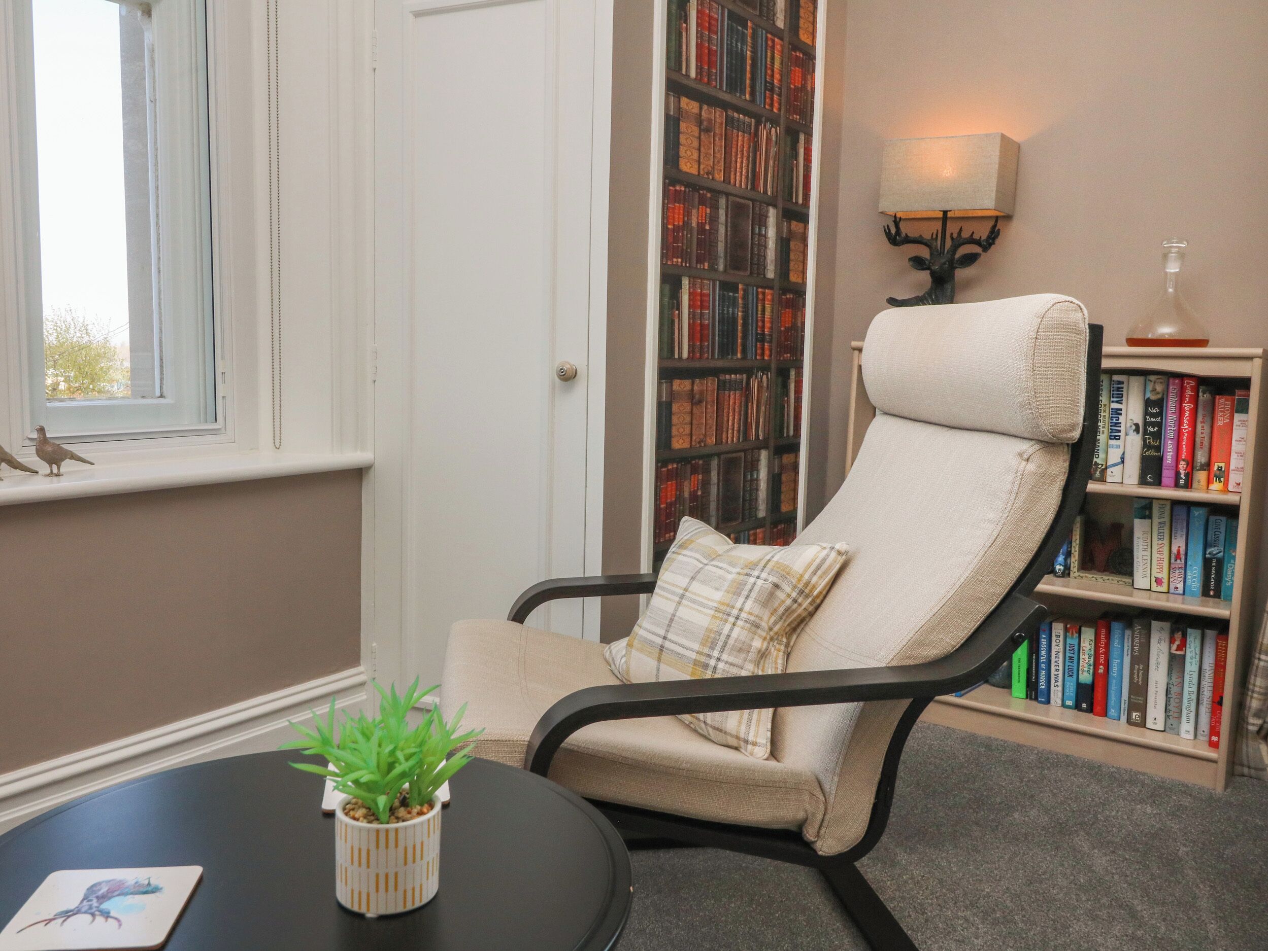A reading chair with a cushion, a bookshelf filled with books, a small table with a plant, and a window with a view.