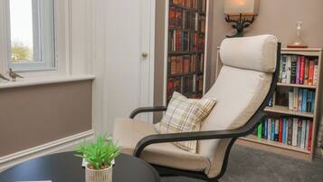 A reading chair with a cushion, a bookshelf filled with books, a small table with a plant, and a window with a view.