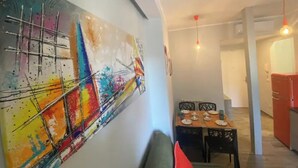 Een moderne woonkamer met een kleurrijk abstract schilderij aan de muur, een eethoek met een tafel gedekt voor vier personen, en een rode koelkast op de achtergrond.