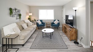Un salon avec un canapé beige, deux fauteuils bleus, une table basse, un téléviseur posé sur un meuble en bois et un tapis à motifs.