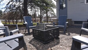 Un espace patio avec un foyer entouré de chaises, une maison en arrière-plan et un grand arbre.