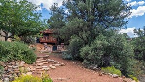 Una baita rustica con veranda in legno e vialetto in pietra, circondata da fitta vegetazione e alberi.