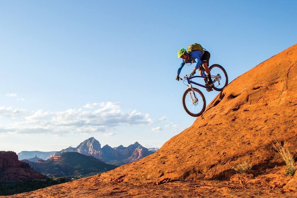 A mountain biker on a steep, rocky terrain with a clear sky and distant mountains in the background.