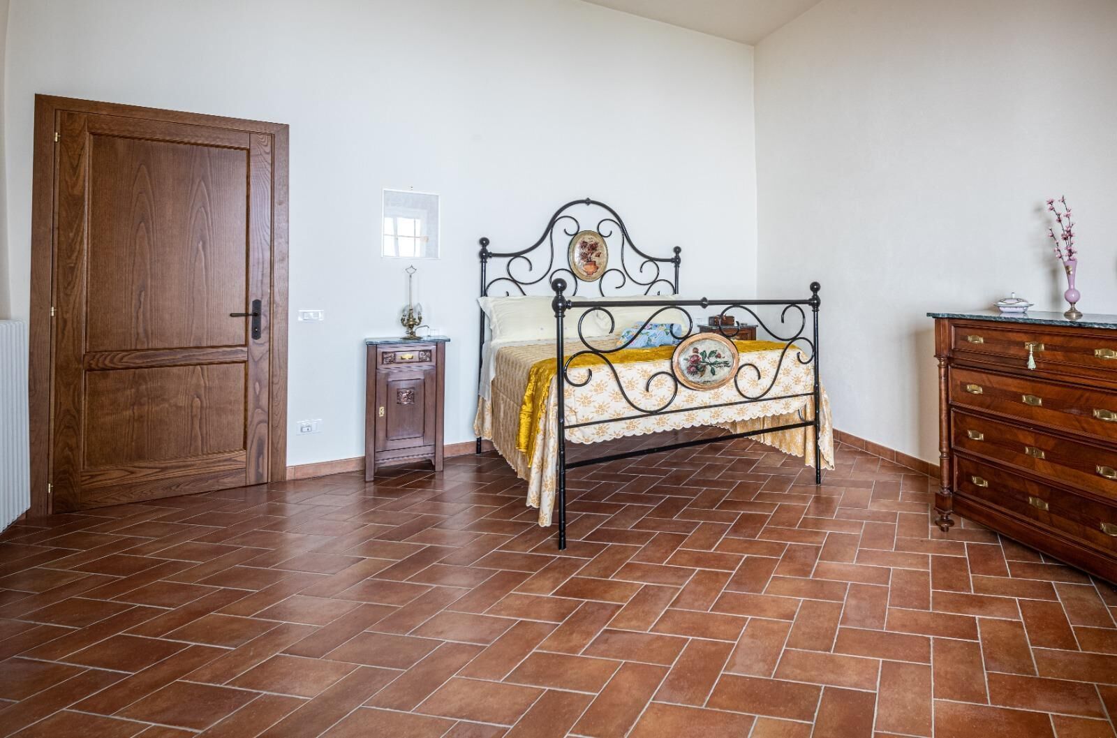 Un dormitorio con puerta de madera, cama con estructura metálica, mesita de noche, cómoda y suelo de baldosas.