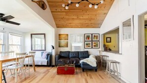 A spacious living area with a wooden ceiling, a dark-colored sofa, a red trunk, and a red rug. - Lakefront Designer House in Pine Plains, NY. (Pine Plains)