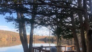 A wooden deck overlooking a calm lake, surrounded by trees. - Lakefront Designer House in Pine Plains, NY. (Pine Plains)