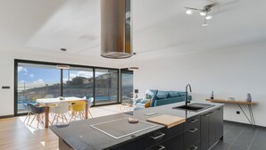A modern kitchen with a central island, a dining area with chairs, a sofa, and a view of the outdoors. - Casa Mozart II (Estreito da Calheta)
