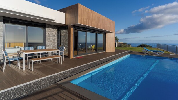 A modern outdoor pool area with a wooden deck, a dining set, and a view of the ocean. - Casa Mozart II (Estreito da Calheta)