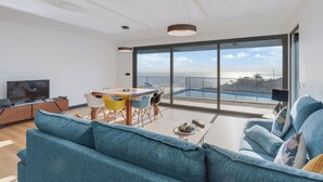 A modern living room with a large sofa, a dining table with chairs, a TV, and a balcony with a pool view. - Casa Mozart II (Estreito da Calheta)