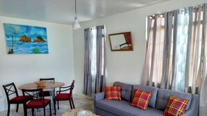 A living room with a gray sofa, red chairs, a small round table, a painting on the wall, and a window with curtains. - Comfortable, local home with A/C and private verandah (Calibishie)