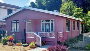 A small, single-story house with a pink exterior, a front porch, and a small garden area with potted plants. - Comfortable, local home with A/C and private verandah (Calibishie)