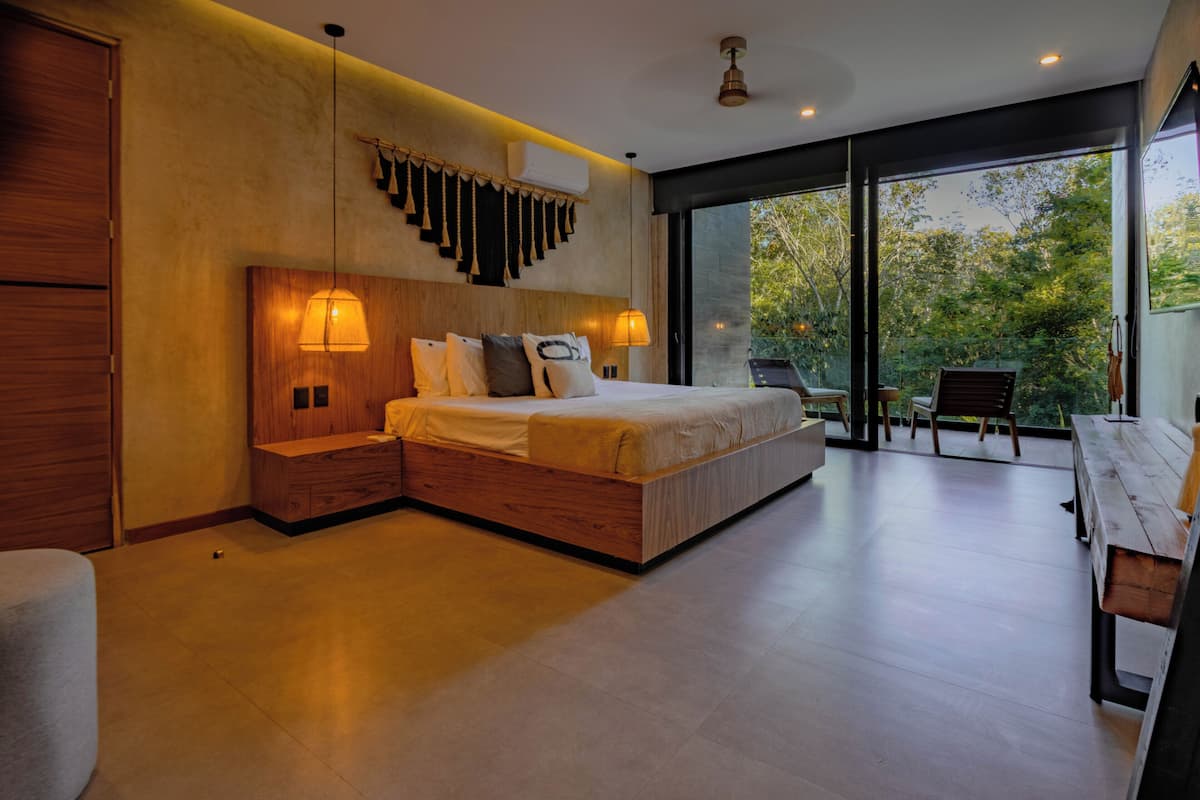 A modern bedroom with a large bed, bedside tables, a wooden bench, and a view of greenery through large windows.