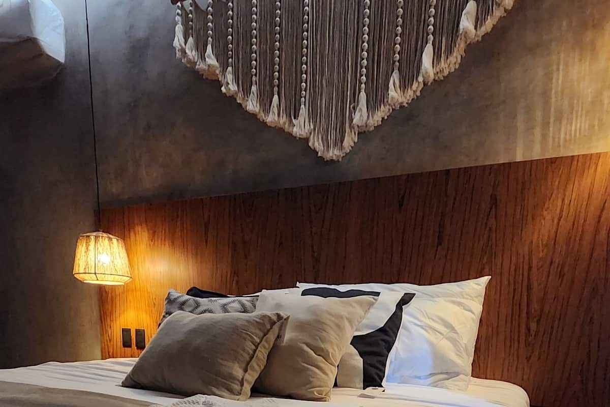 A modern bedroom with a wooden headboard, a large bed with multiple pillows, a textured throw blanket, and a hanging macramé wall decoration.