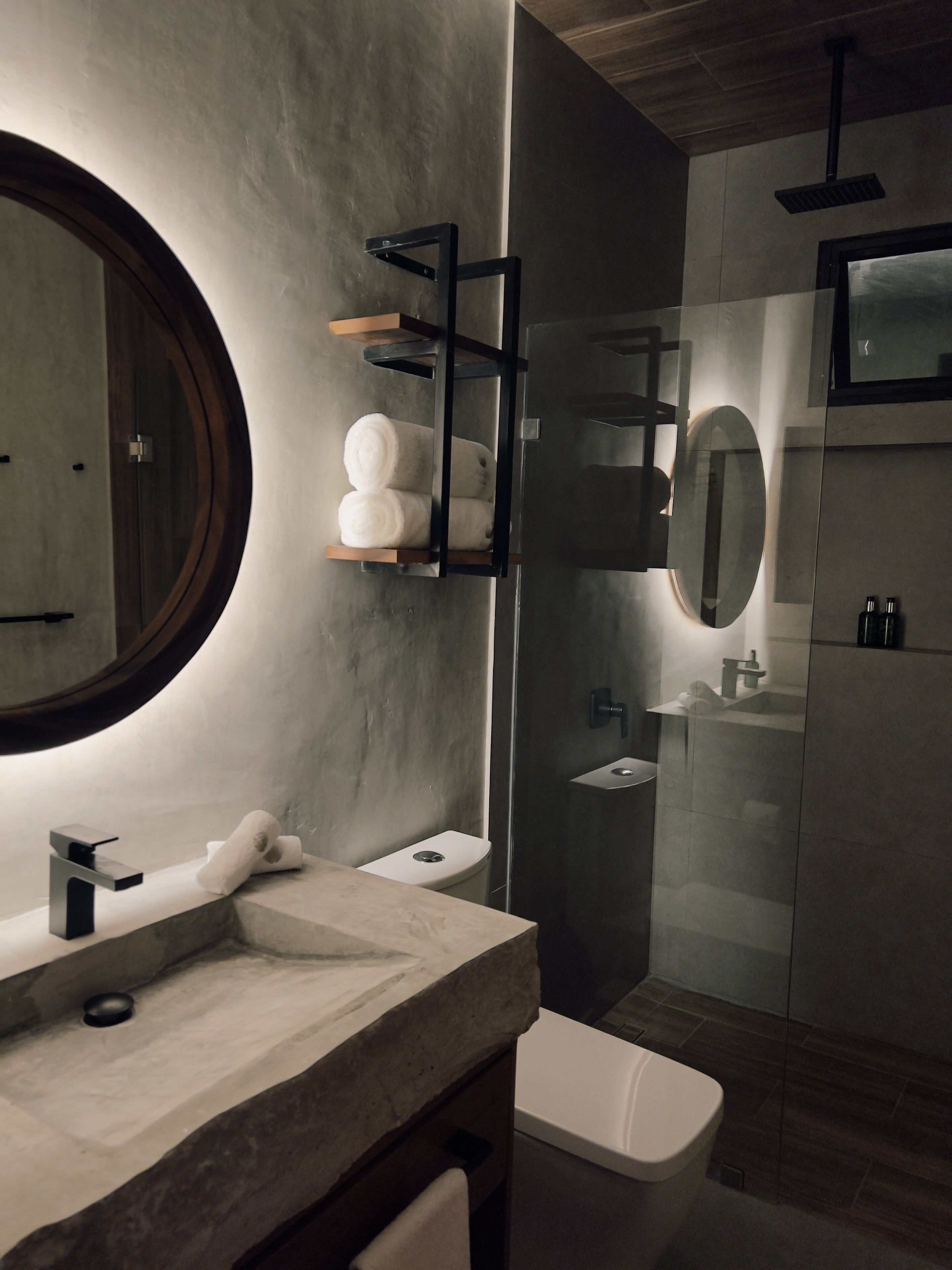 A modern bathroom with a round mirror, a marble sink, a toilet, and a glass-enclosed shower area.