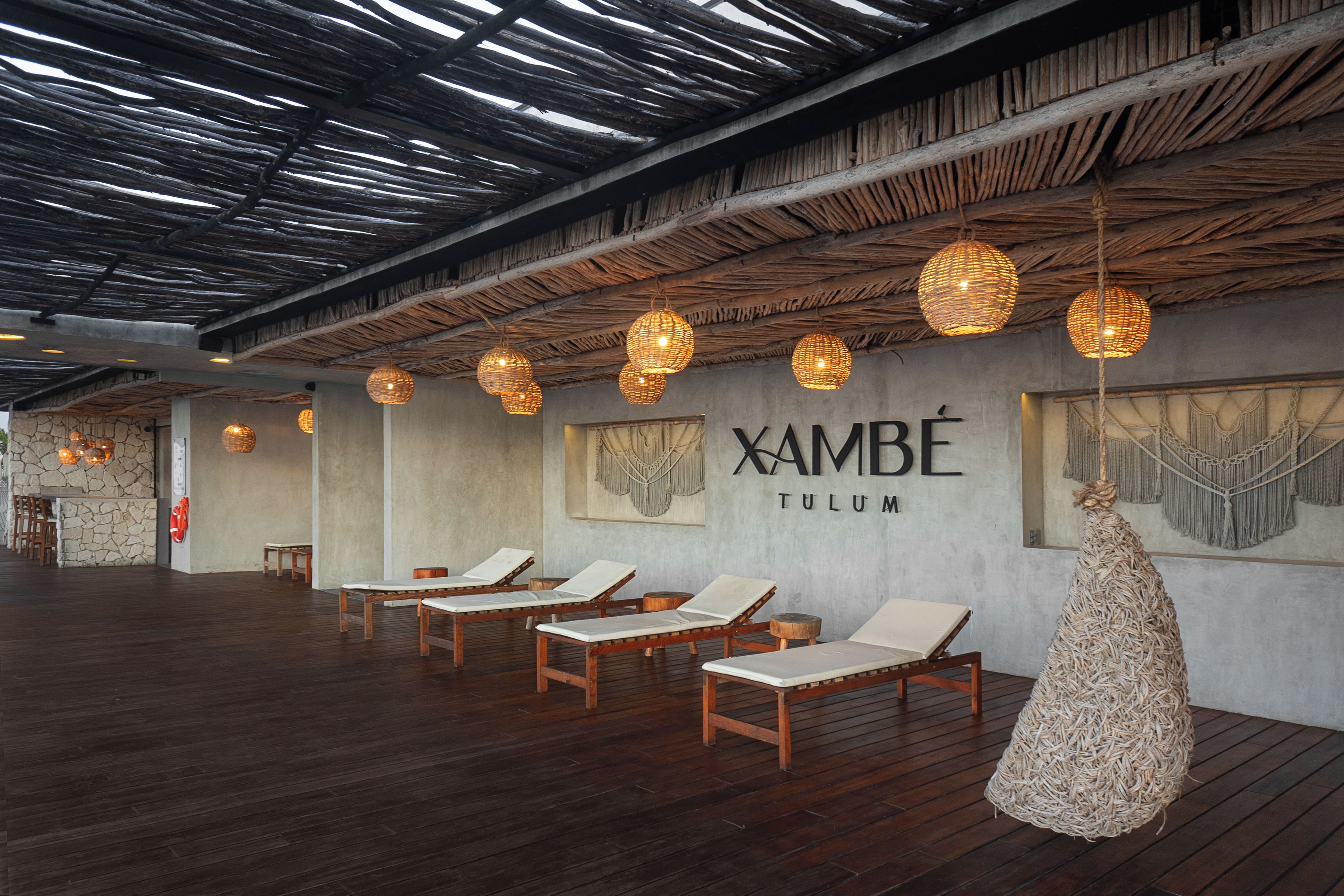 A spa area with wooden loungers, woven light fixtures, and a wall with the name 'XAMBE TULUM'.