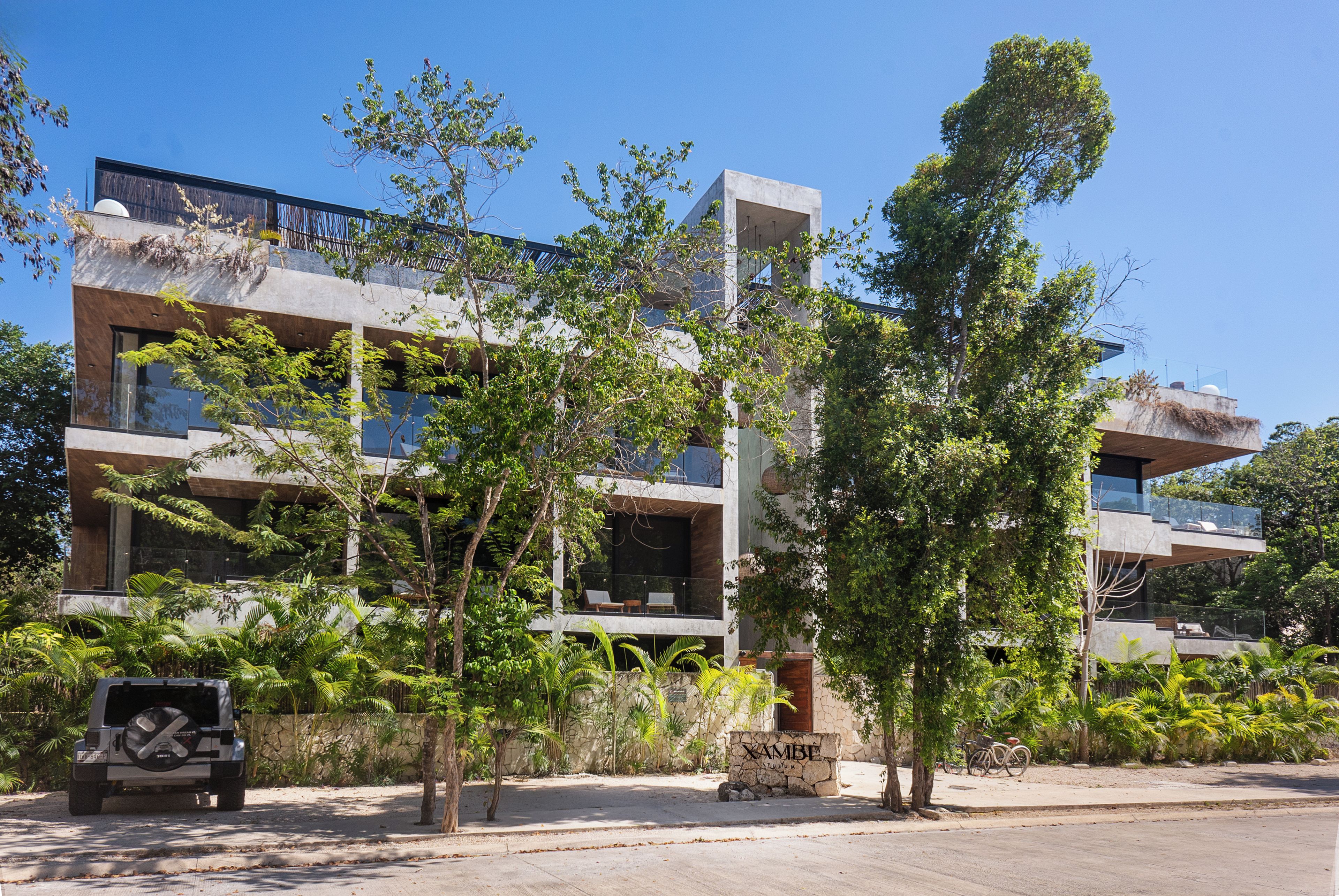 A modern building with multiple balconies, surrounded by lush greenery and a clear sky.