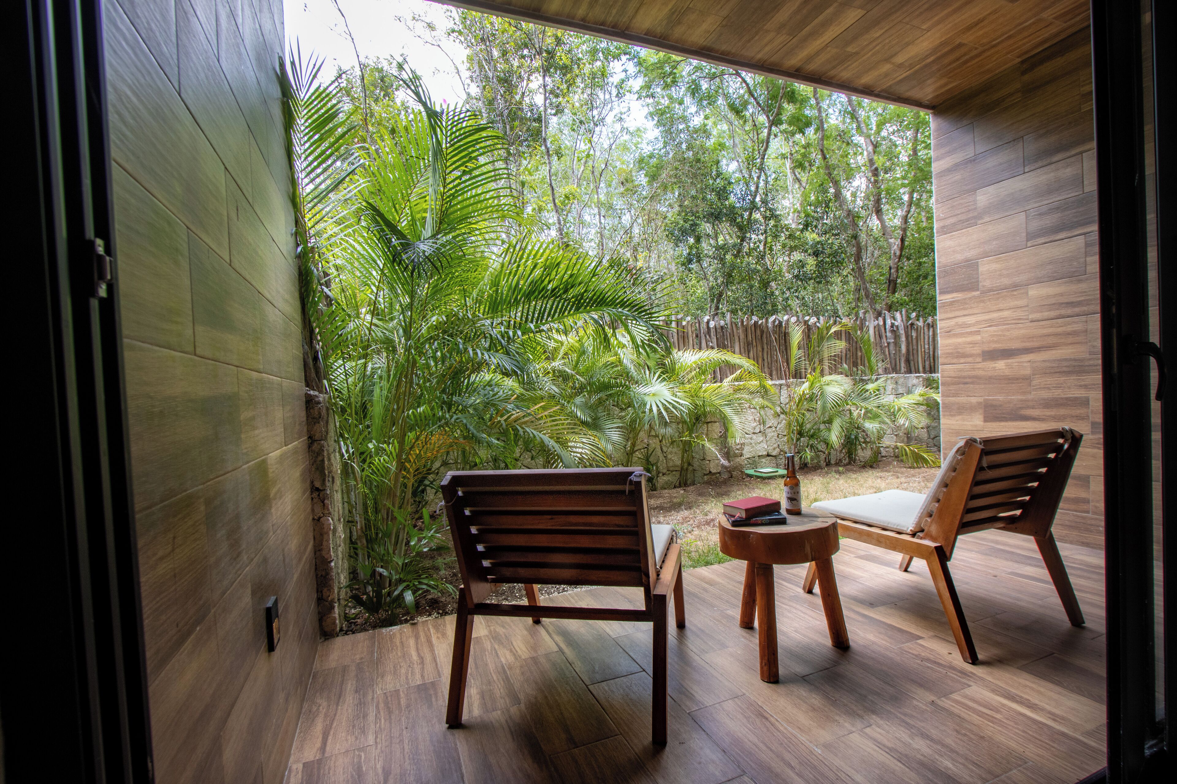 An outdoor patio with wooden flooring, two wooden chairs, a small round table, and lush greenery in the background.