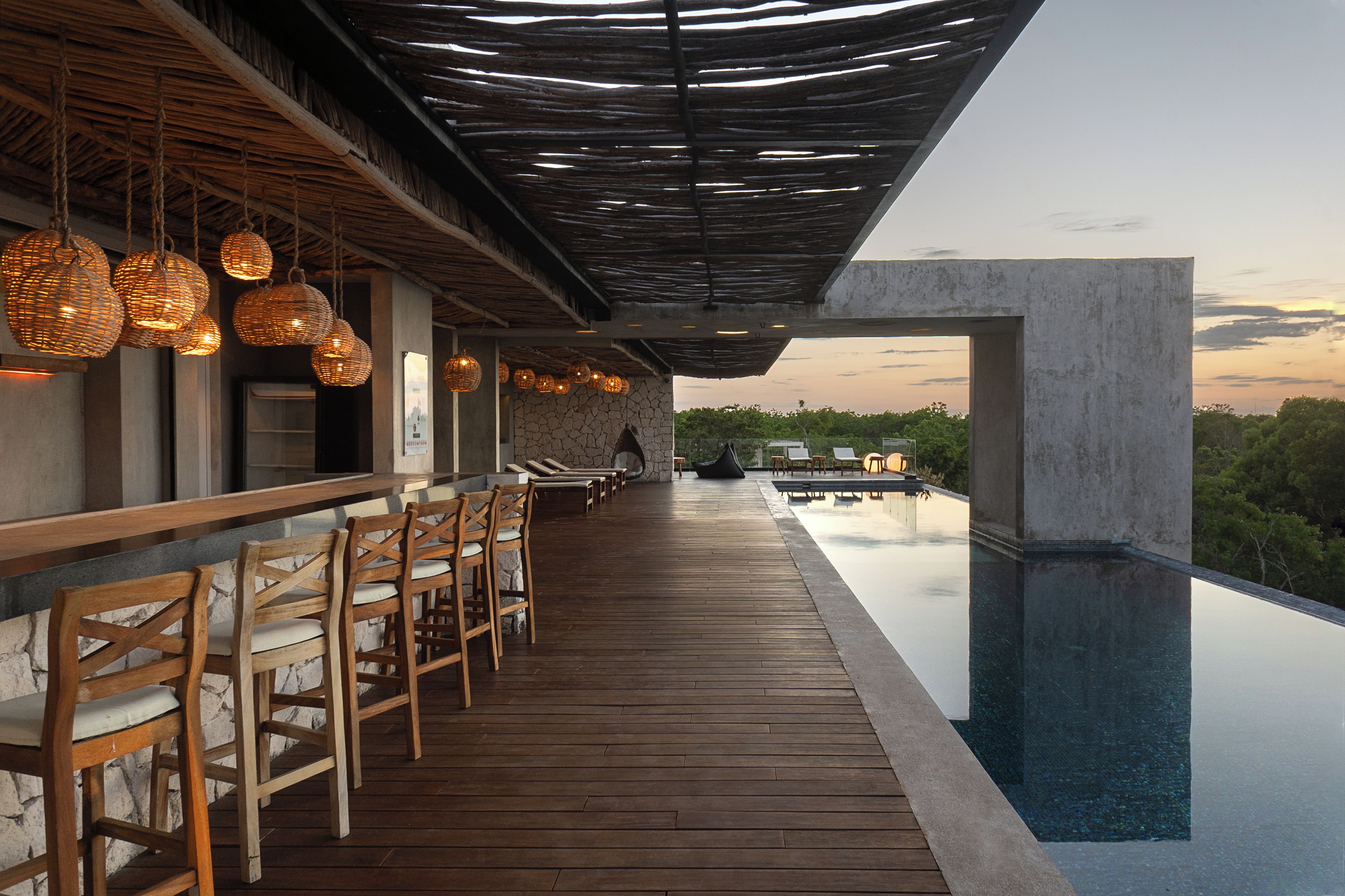 A bar area with wooden chairs and hanging wicker lamps, overlooking a pool.