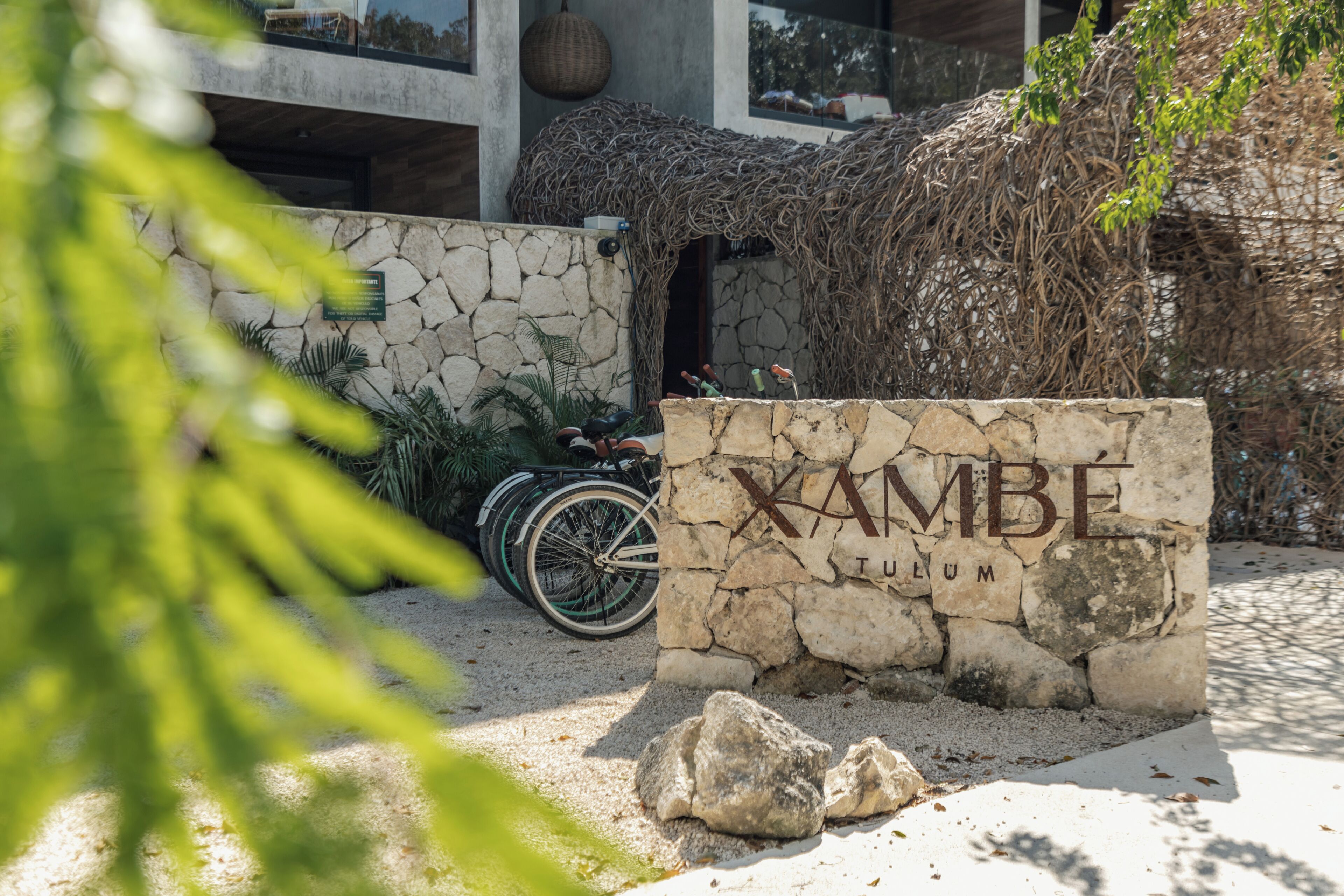 A stone sign with 'XAMBE' and 'TULUM' inscribed, a bicycle, and a stone wall with greenery.