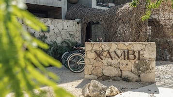 A stone sign with 'XAMBE' and 'TULUM' inscribed, a bicycle, and a stone wall with greenery.