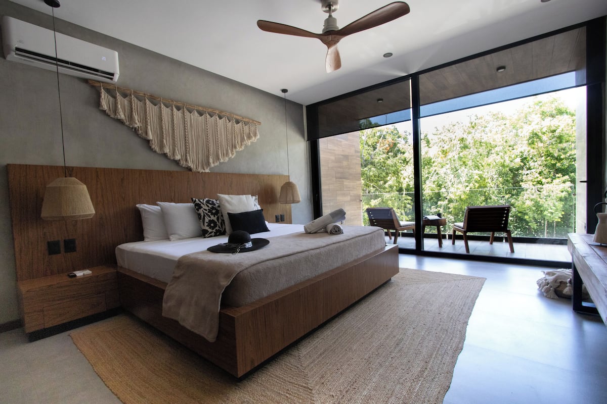 A modern bedroom with a large bed, a ceiling fan, a view of the outdoors, and a wooden headboard.