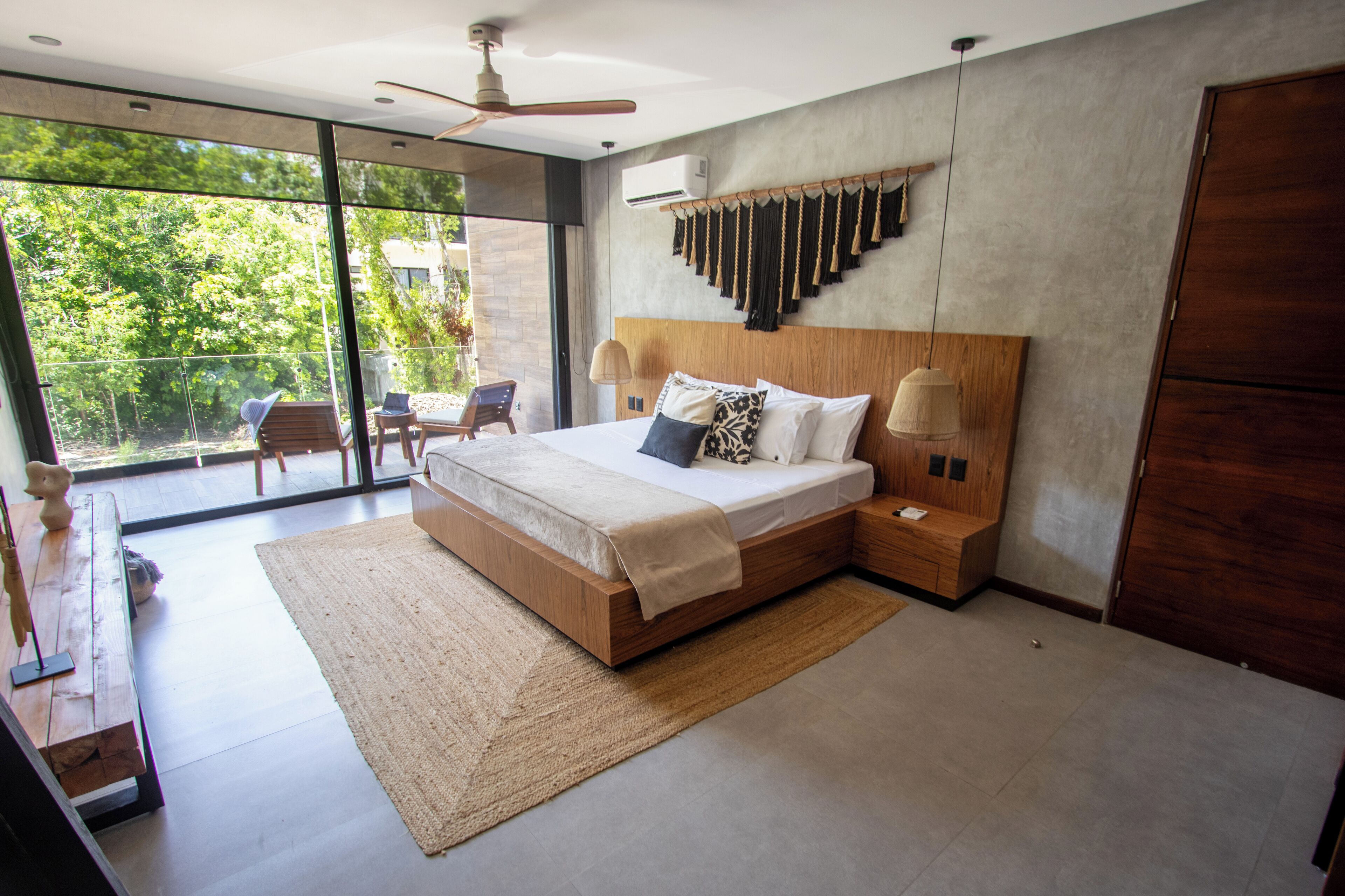 A modern bedroom with a large bed, wooden headboard, and a view of greenery through large windows.
