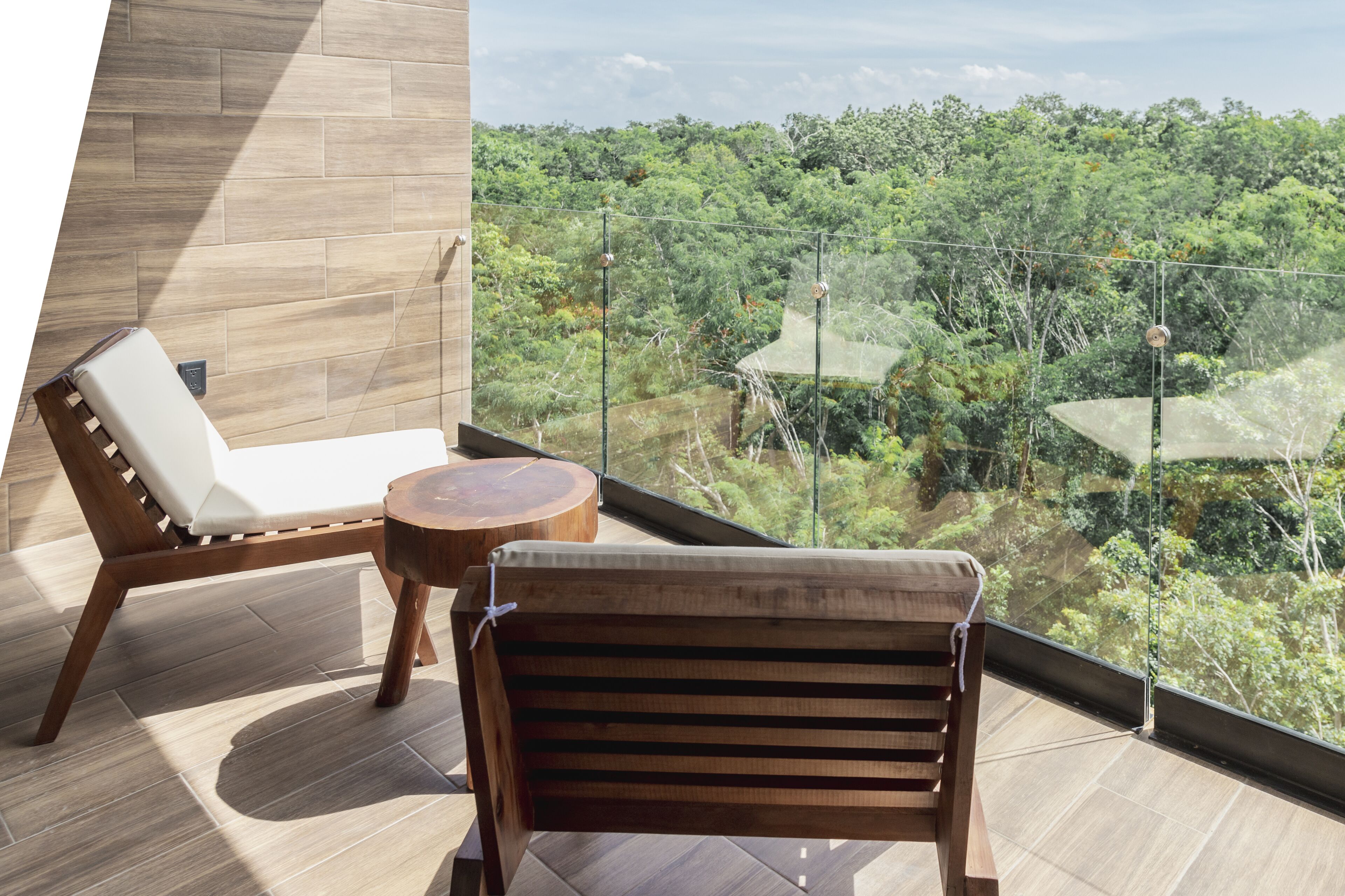 Un balcón con una silla de madera y una pequeña mesa redonda, con vistas a un bosque frondoso y verde.