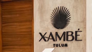 Un mostrador de recepción con encimera de madera, un cartel en la pared que dice "XAMBÉ TULUM" y una lámpara colgante.