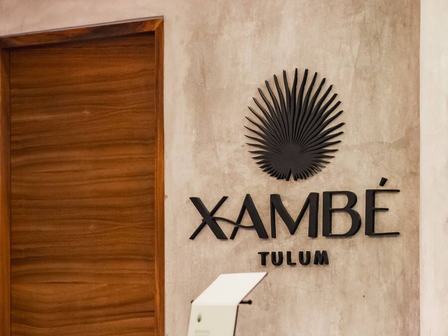 Un mostrador de recepción con encimera de madera, un cartel en la pared que dice "XAMBÉ TULUM" y una lámpara colgante.