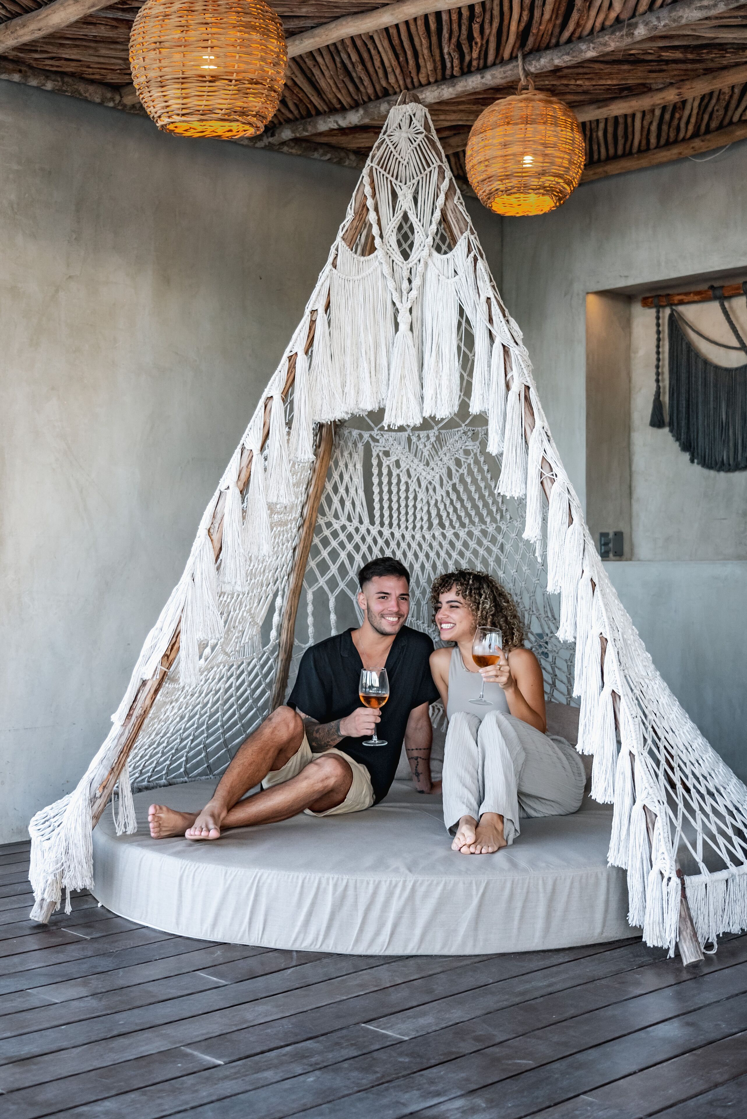 A couple sitting in a macrame hammock with a concrete floor and wooden ceiling.