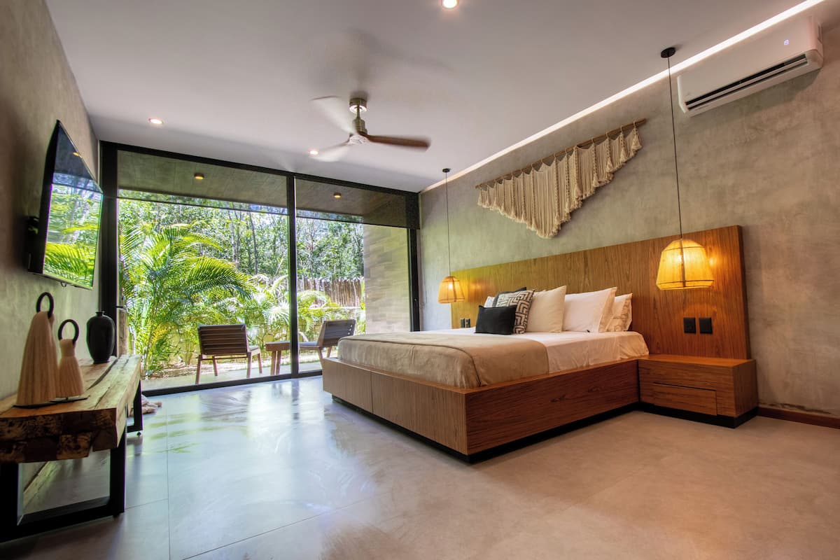 A modern bedroom with a large bed, a ceiling fan, and a view of lush greenery through large windows.