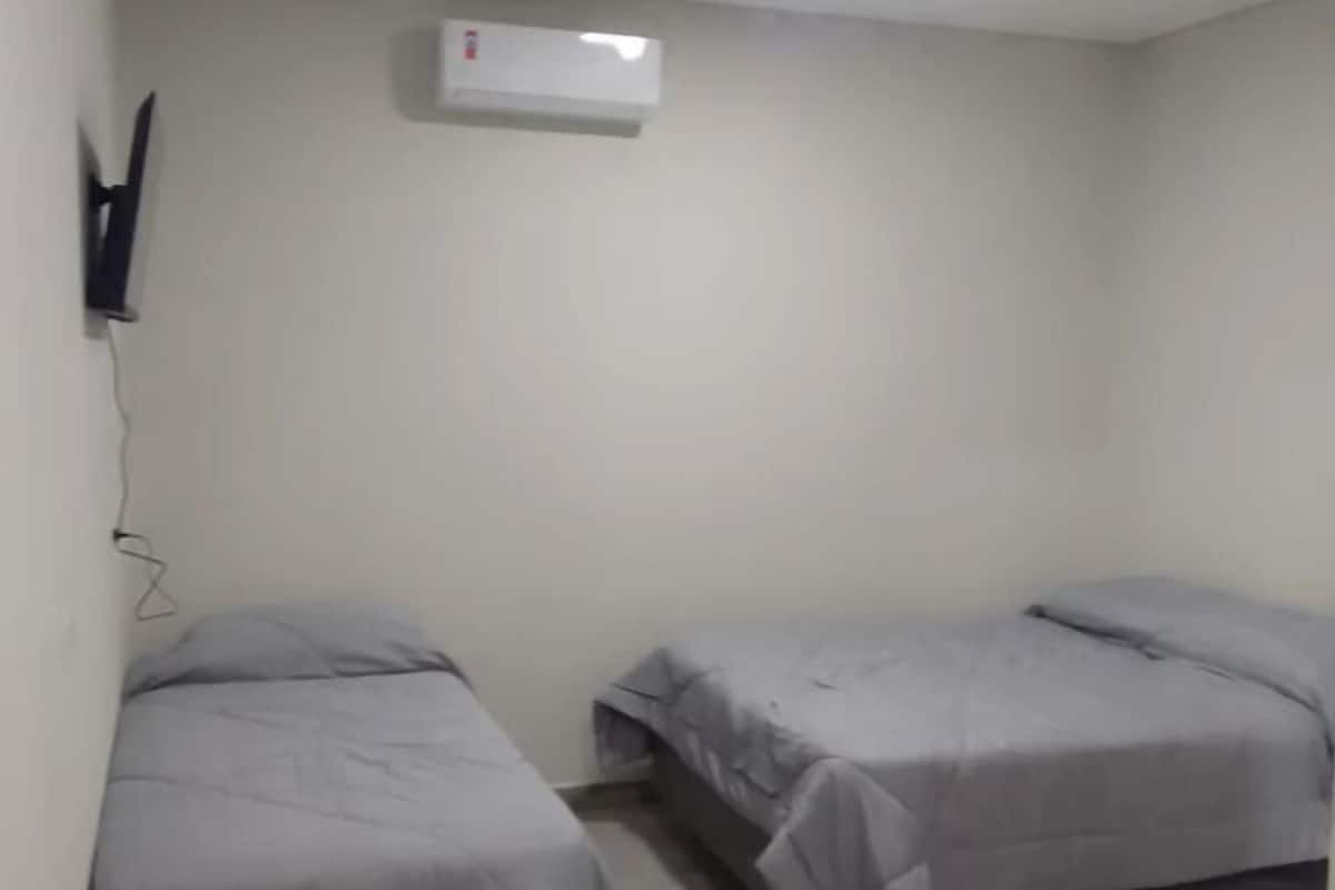 Una habitación pequeña y limpia con dos camas individuales, aire acondicionado instalado en la pared y un televisor a la izquierda.