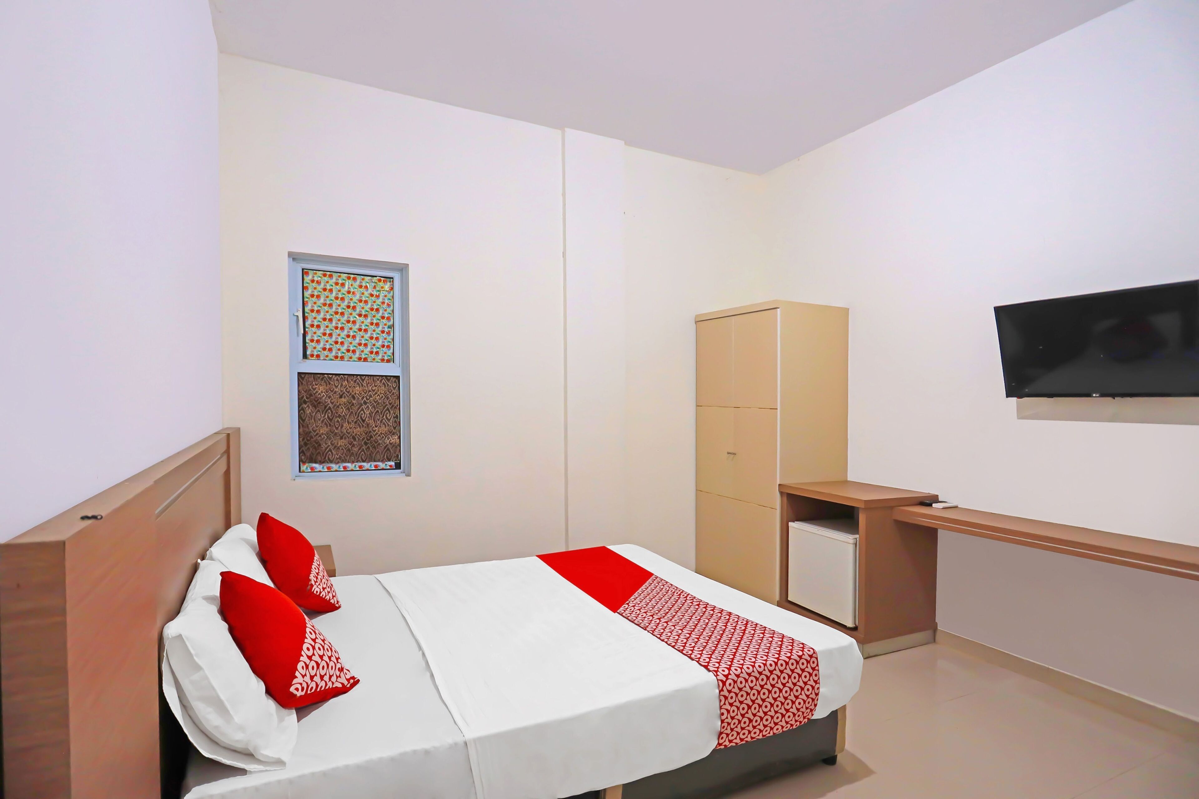 A bedroom with a bed, a wardrobe, a desk, a television, and a window with patterned curtains.