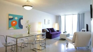 Une chambre d’hôtel moderne avec une table à manger en verre, un canapé violet, un fauteuil blanc et un téléviseur fixé au mur.