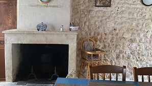 A rustic interior with a stone wall, a fireplace, wooden beams, a clock, and a dining area with chairs and a table.