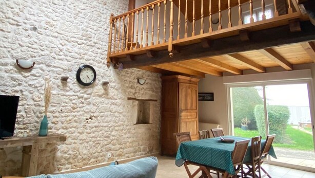 A rustic interior with a stone wall, wooden ceiling beams, a loft area with a wooden railing, a dining table with chairs, and a sofa.