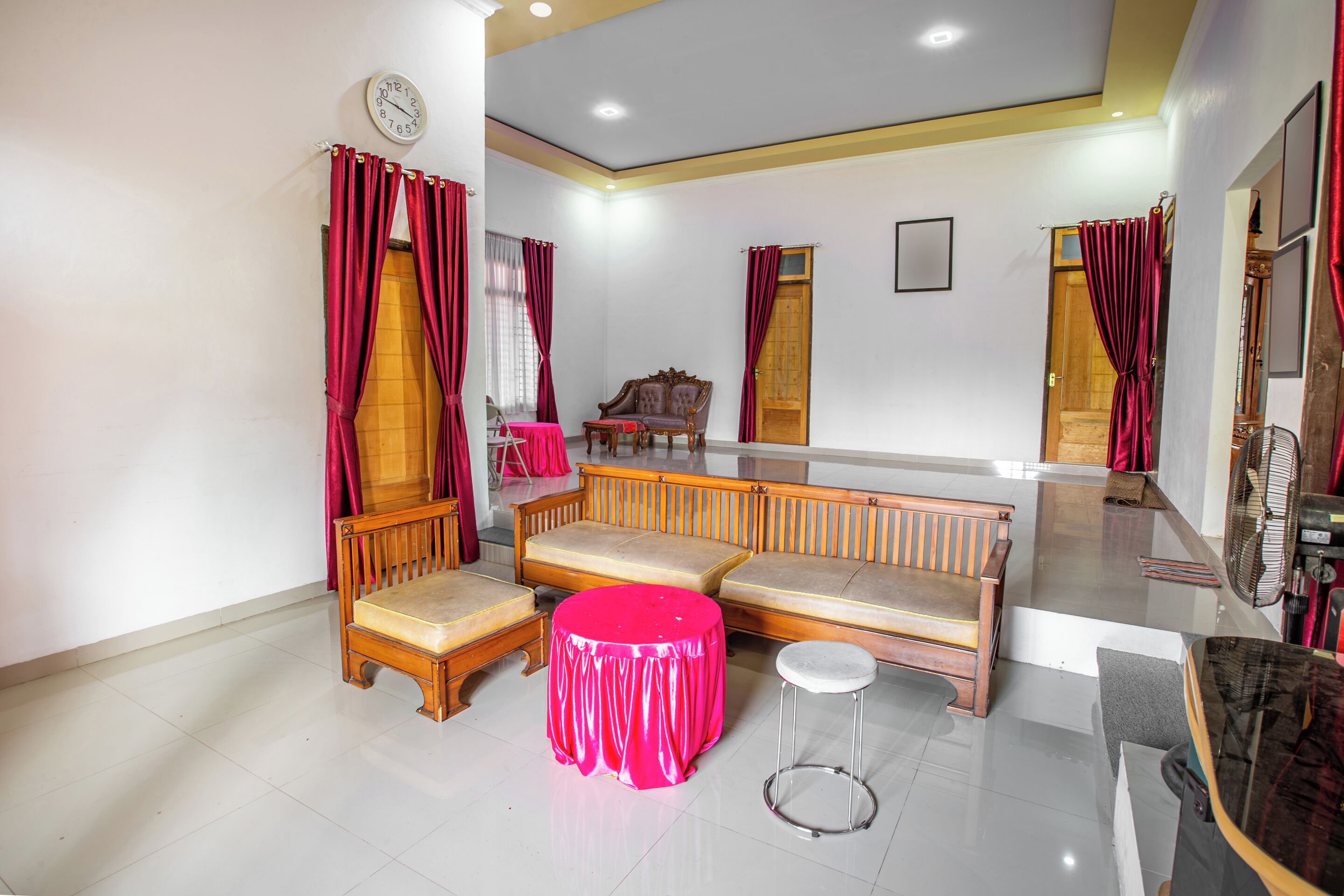 A room with a wooden sofa, a round table with a pink tablecloth, and a small stool. There is a wooden bench, a mirror, and a fan on the wall.