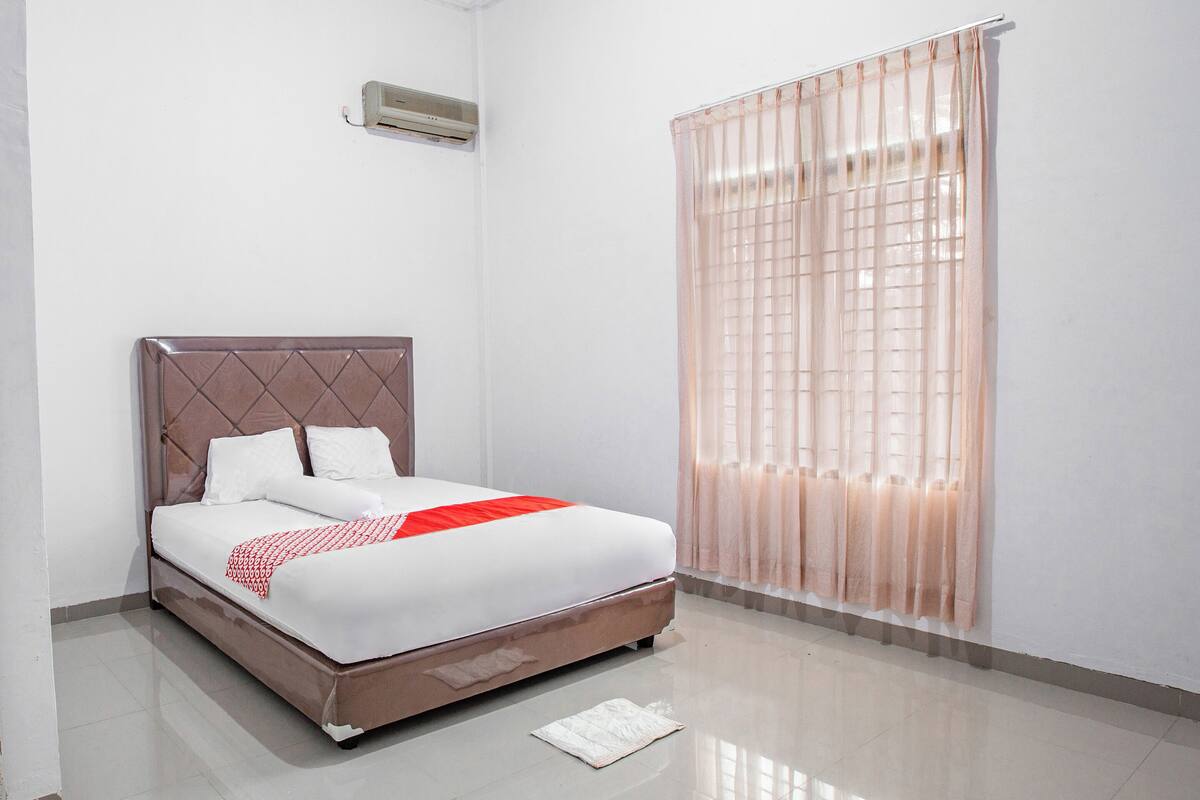A neatly made bed with a brown headboard, a white and red bedspread, and a window with sheer curtains.