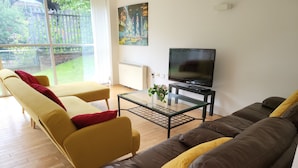 A living room with a brown leather sofa, yellow armchairs, a glass coffee table, and a flat-screen TV. - Luxury River View 2-bed Apt, Wifi, Garden, River (Salford)