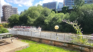 A riverside walkway with a boat tour, surrounded by greenery and urban buildings. - Luxury River View 2-bed Apt, Wifi, Garden, River (Salford)