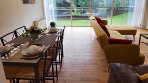 A dining area with a table set for four, a sofa, and a large sliding glass door leading to a garden. - Luxury River View 2-bed Apt, Wifi, Garden, River (Salford)