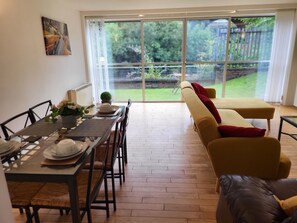 A dining area with a table set for four, a sofa, and a large sliding glass door leading to a garden. - Luxury River View 2-bed Apt, Wifi, Garden, River (Salford)