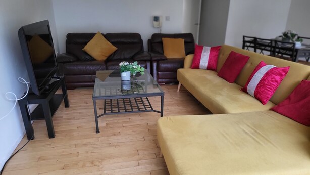 A living room with a yellow sofa, a glass coffee table, a brown leather couch, and a television on a stand. - Luxury River View 2-bed Apt, Wifi, Garden, River (Salford)