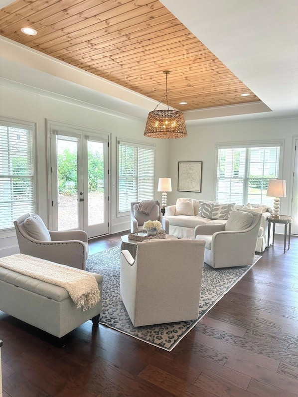 A spacious living room with a high ceiling, large windows, and a central chandelier.